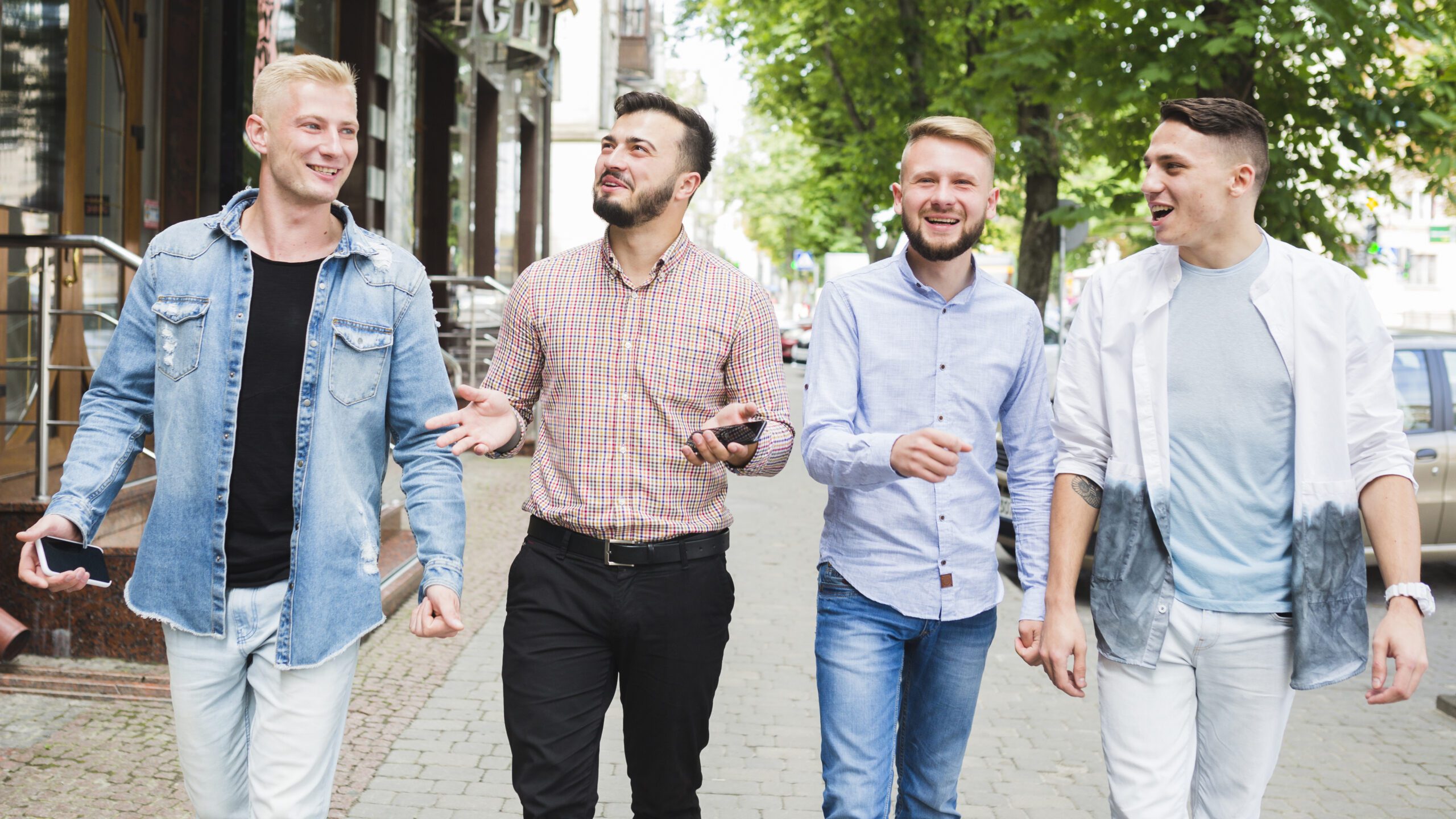 man-holding-mobile-phone-his-hand-enjoying-with-his-friend-street Four men walking and talking on sidewalk.